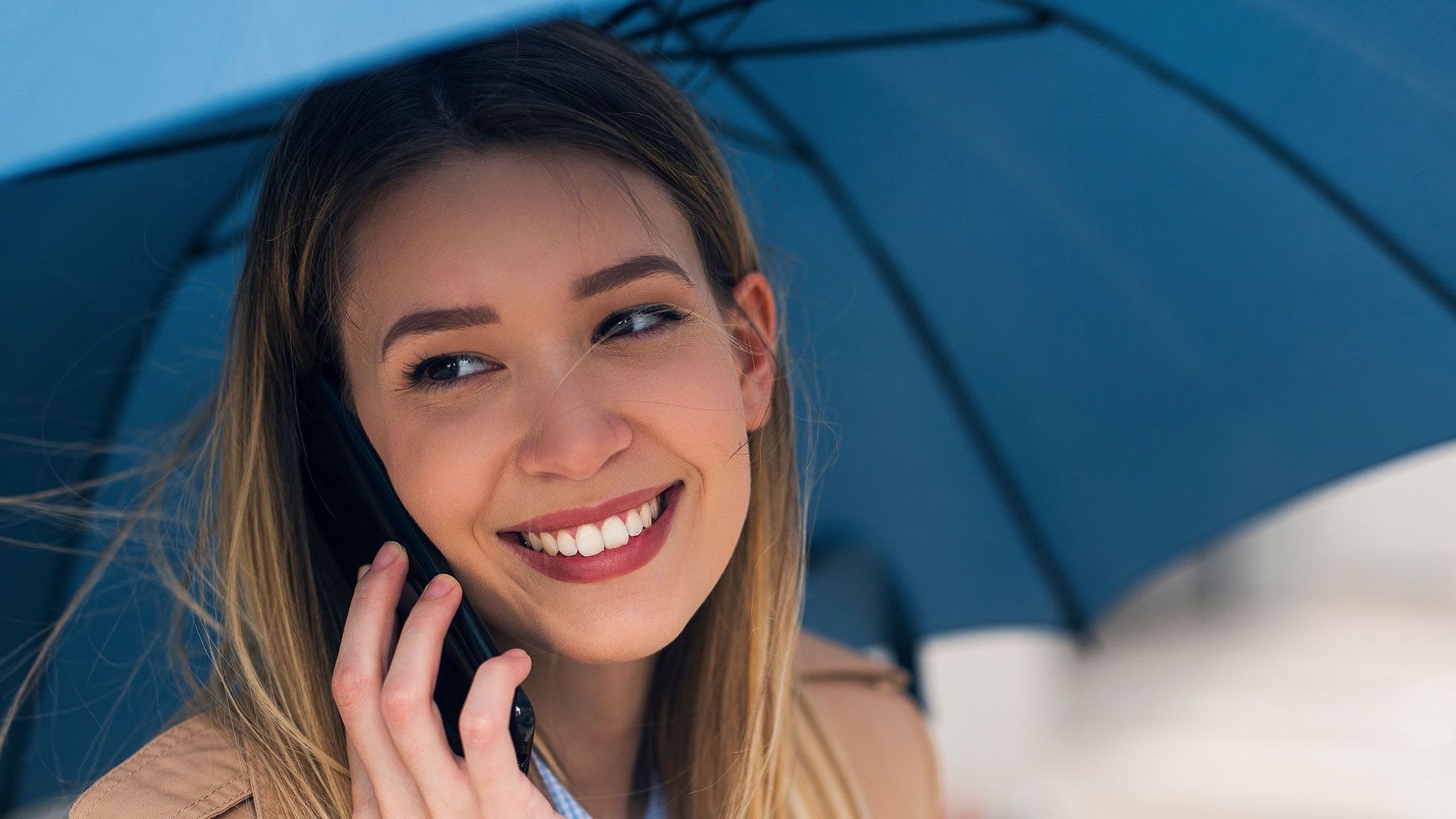 An image of a lady smiling while holding an umbrella while on the phone